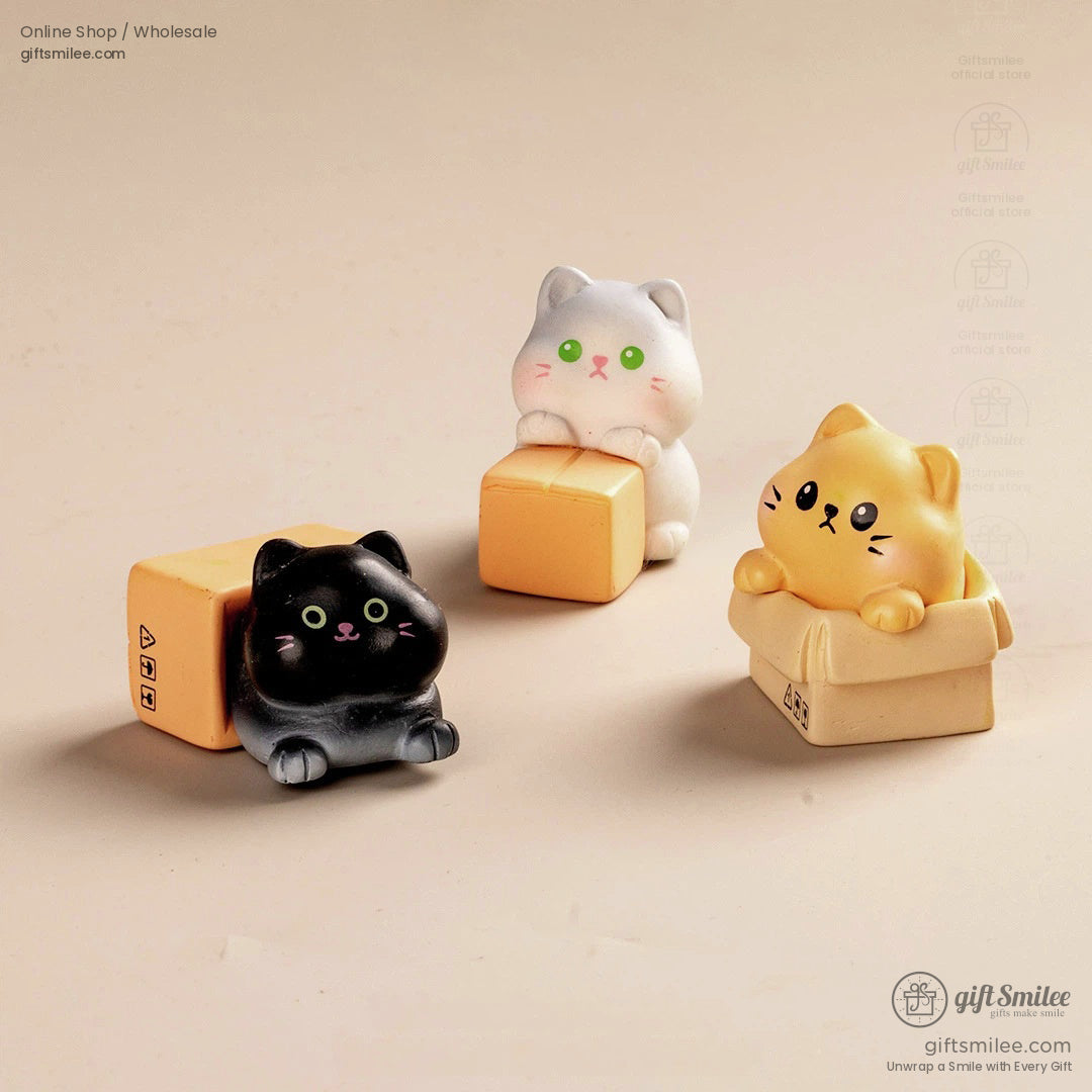 Three cat figurines in black white and yellow with green eyes paired with small cardboard box props on a beige surface