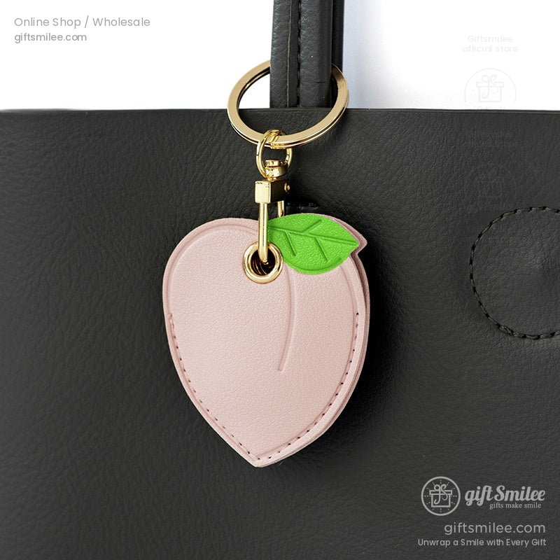Pastel pink faux leather peachshaped keychain with a green leaf detail and gold metal clasp hanging on a black bag