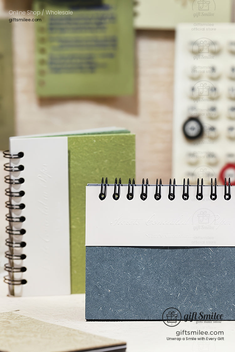 Minimalist spiral-bound desk notebooks in green, white, and blue covers displayed on a workspace from giftsmilee.com