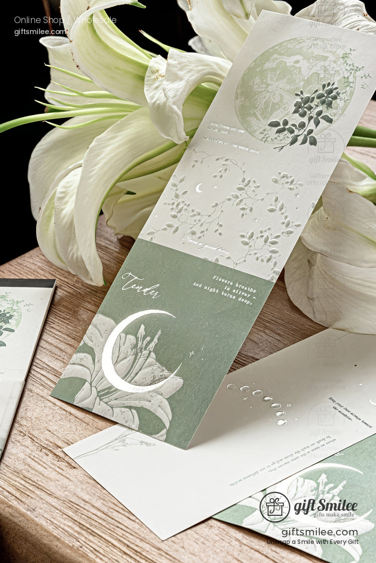 Elegant green and white floral-themed greeting card with embossed moon and leaves, displayed on a wooden surface beside white lilies.