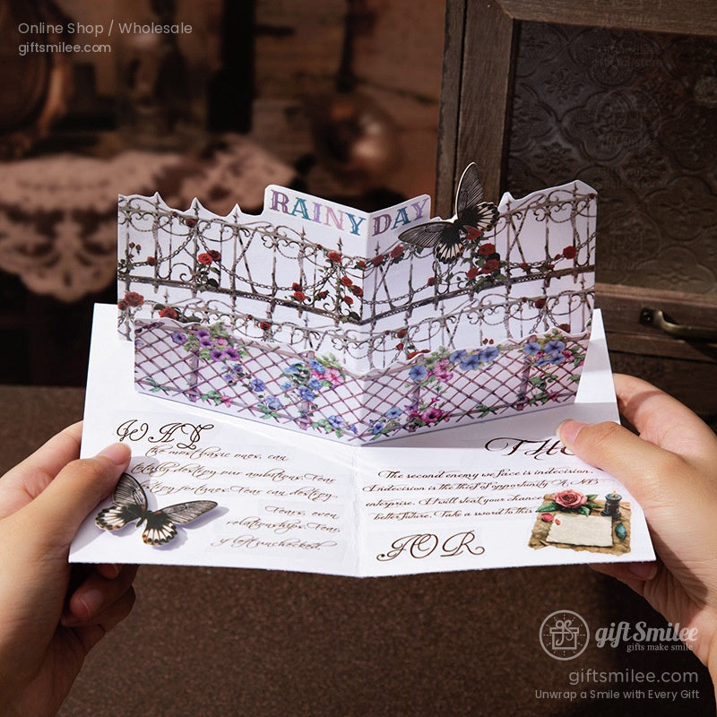 Pop-up “Rainy Day” greeting card with floral fence and butterfly design being held open