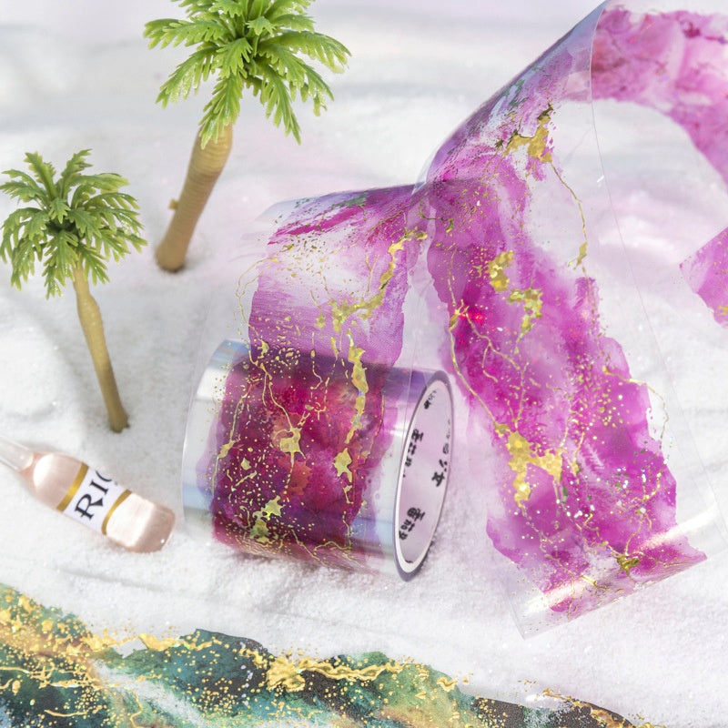 Transparent washi tape with vibrant purple and pink watercolor patterns accented by gold foil streaks on a white backdrop