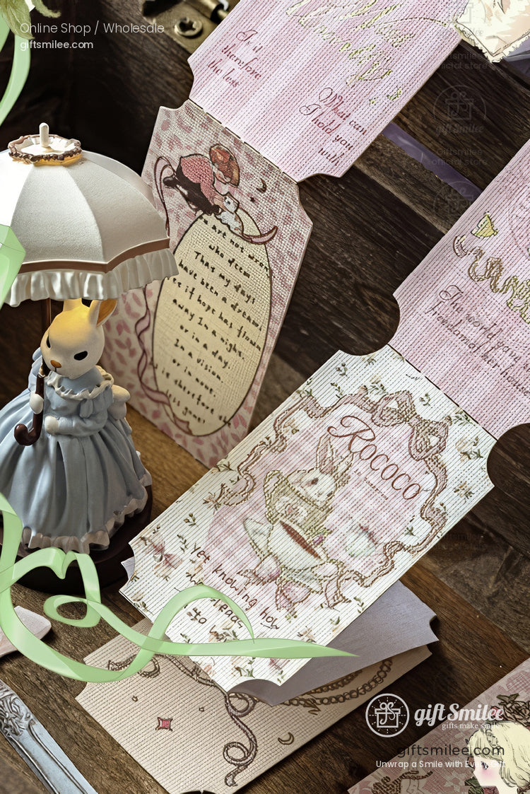 Vintage-style pastel stationery and Rococo-themed decorative tickets displayed with a small figurine and ribbon on a wooden desk