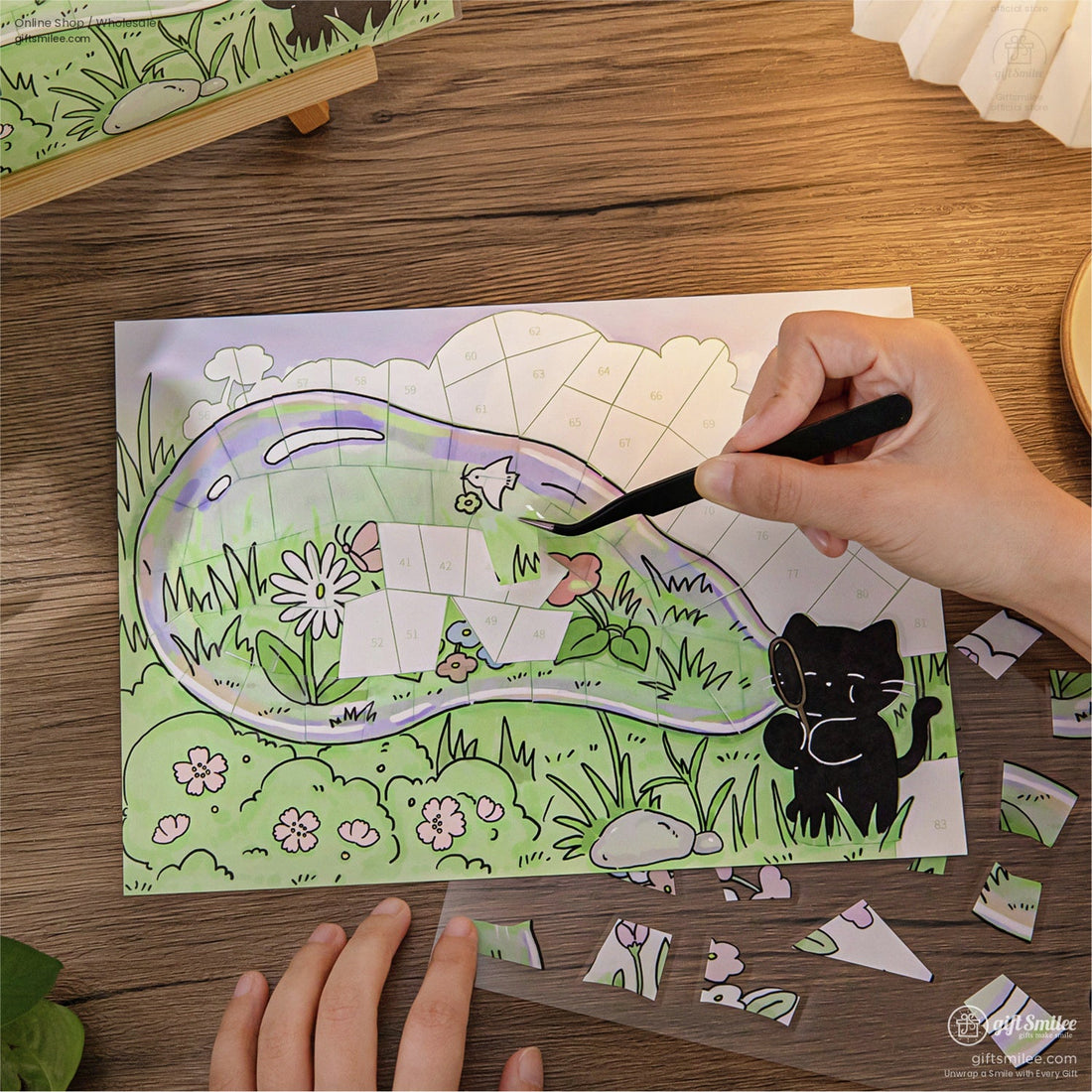 Person assembling a cute cat-themed mosaic puzzle on a wooden table, craft tweezers placing numbered pieces in a green garden scene.
