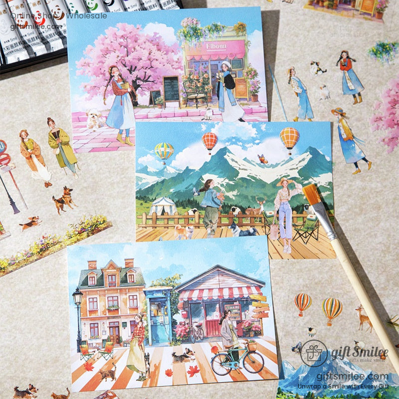 Cute watercolor travel postcards and stickers featuring scenic towns, hot air balloons, mountains, and pedestrians on a creative flat lay with paintbrush – scrapbooking stationery set by Giftsmilee.
