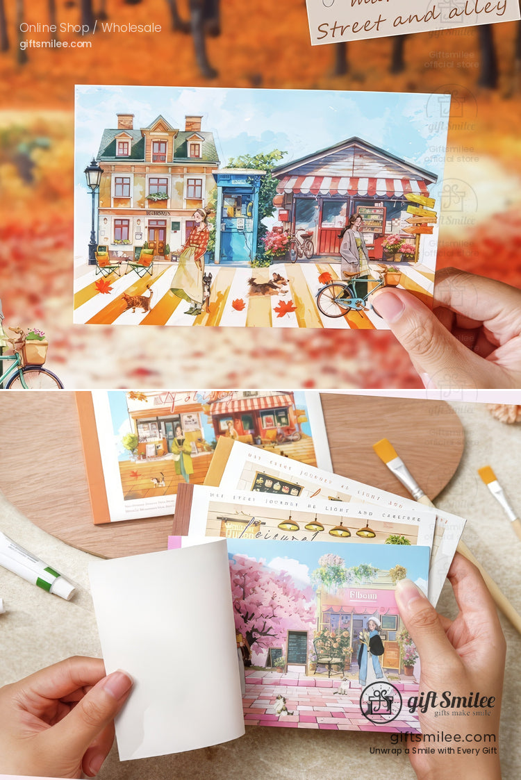 Hand holding a colorful illustrated postcard of a quaint street scene with shops, bicycle, and pedestrians, decorative stationery set for journaling or scrapbooking.