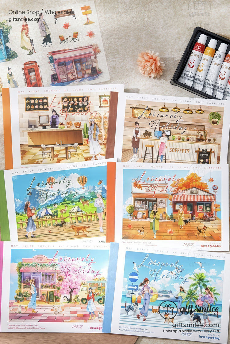 Set of “Heavenly Holiday” illustrated postcards laid out on a desk with paint tubes, showing cozy café, bakery, hot air balloons, and travel scenes from Giftsmilee.