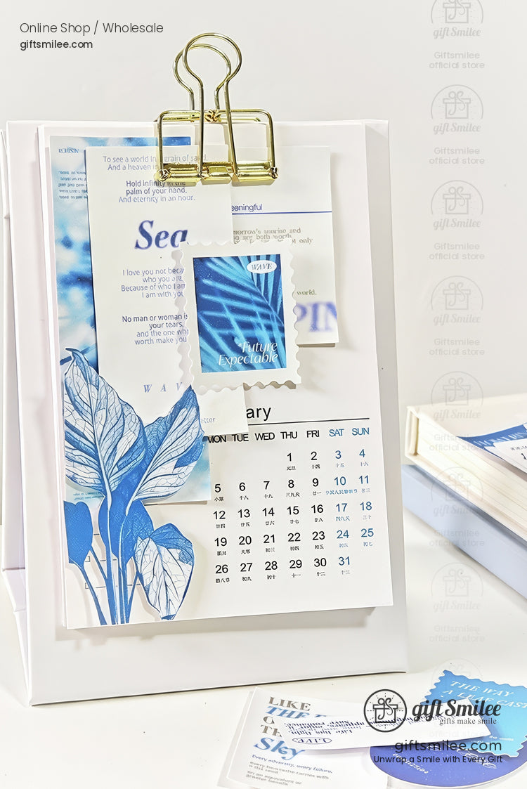 Minimalist white desk calendar with blue botanical prints gold binder clip and layered artistic paper accents